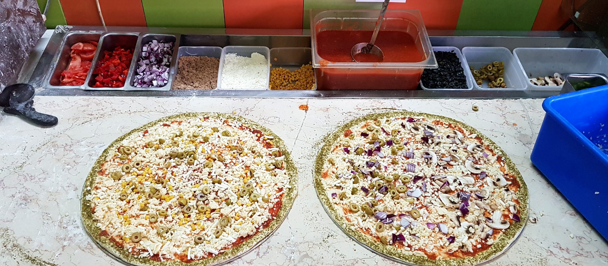שתי פיצות נבנות על דלפק העבודה, מול מגשי חומרי הגלם
